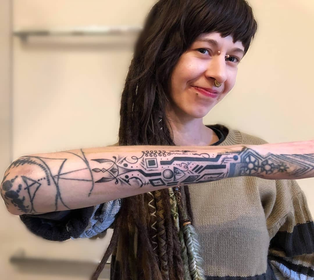 Smiling woman showing a detailed cyberpunk tattoo on her forearm with geometric and circuit-like designs